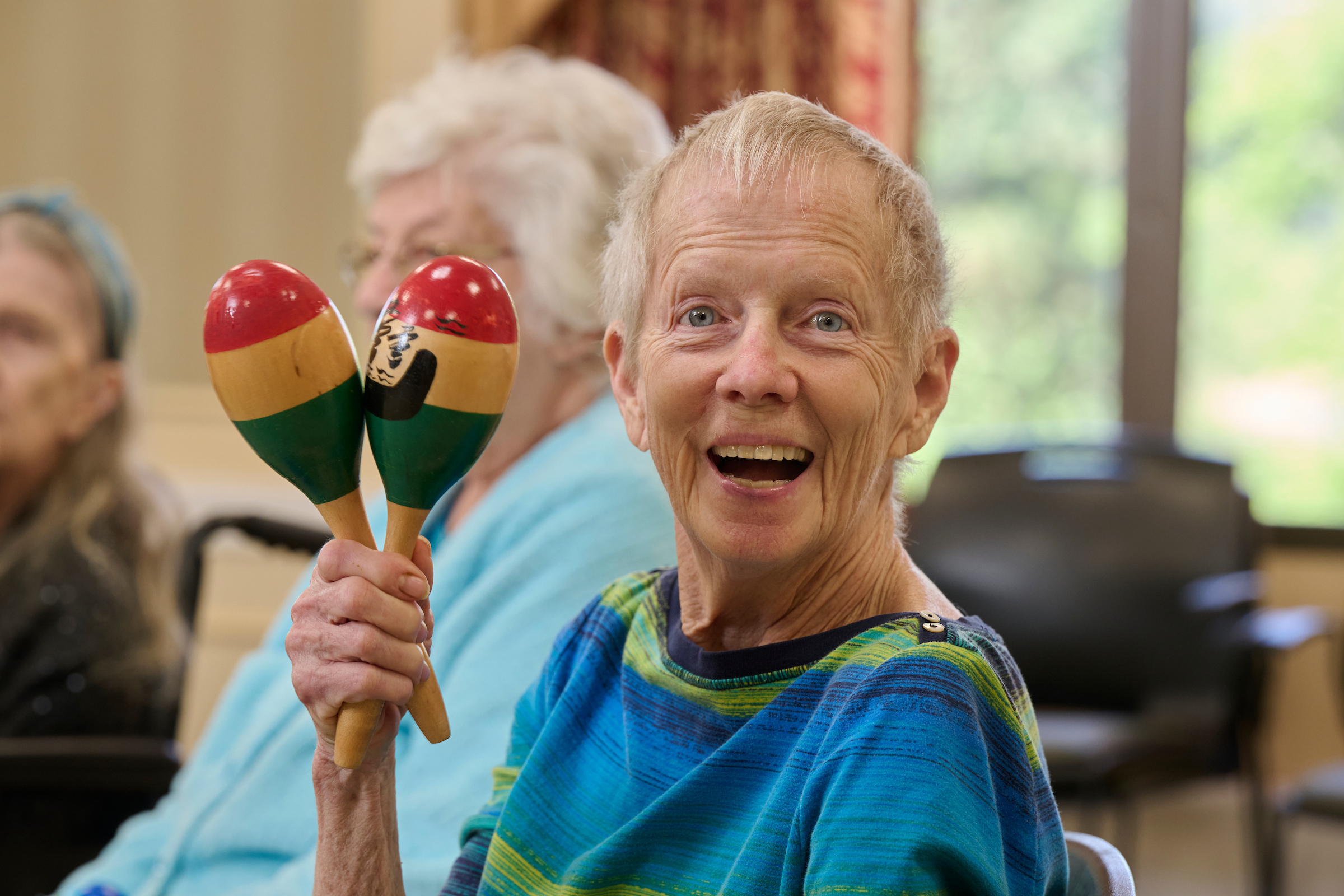 Smiling elderly woman holding colorful maracas during a group activity in a bright room.