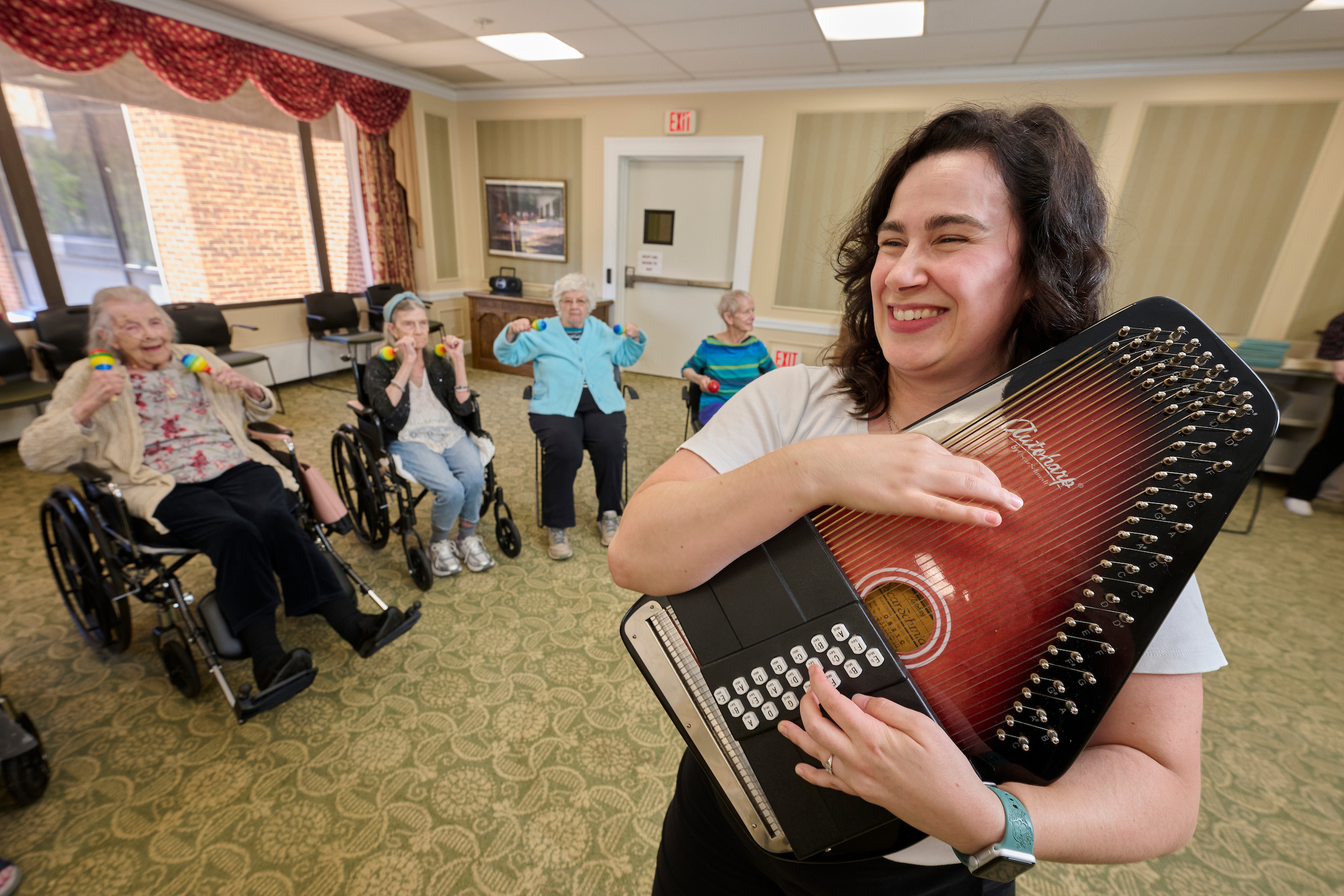 Woman in the foreground smiles while playing a large red guitar-like instrument; elderly residents in wheelchairs watch in a cozy activity room.