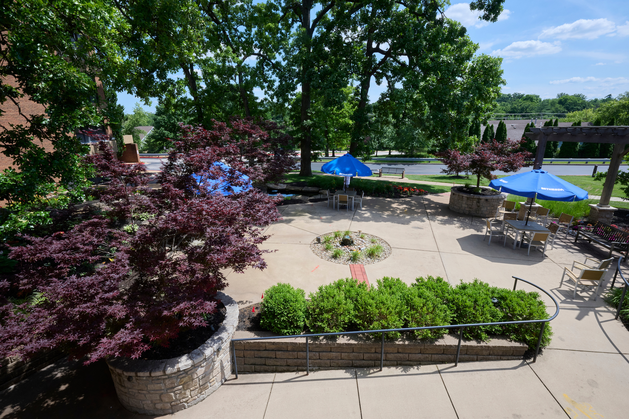 Outdoor patio area with blue Bethesda umbrellas, tables, and chairs surrounded by trees and stone planters.