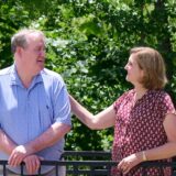 A man and a woman stand outdoors by a railing, smiling at each other; the woman rests her hand on the man's shoulder with green trees in the background.