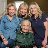 Smiling elderly man seated in a green plaid shirt with three women standing behind him in a cozy family portrait.