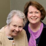 Two women smiling at the camera, standing side by side indoors, posing for a photo.
