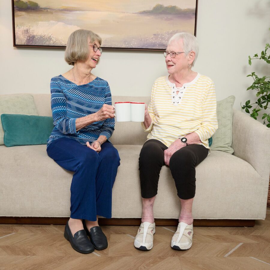 Two smiling elderly women sit on a beige sofa, clinking white mugs with red rims in a cozy living room.