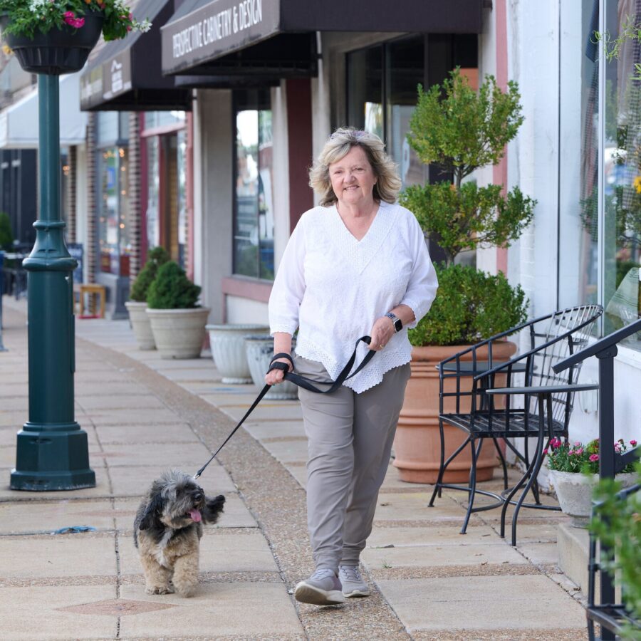 Woman walking a small fluffy dog on a leash along a sidewalk in front of shops with planters and seating.
