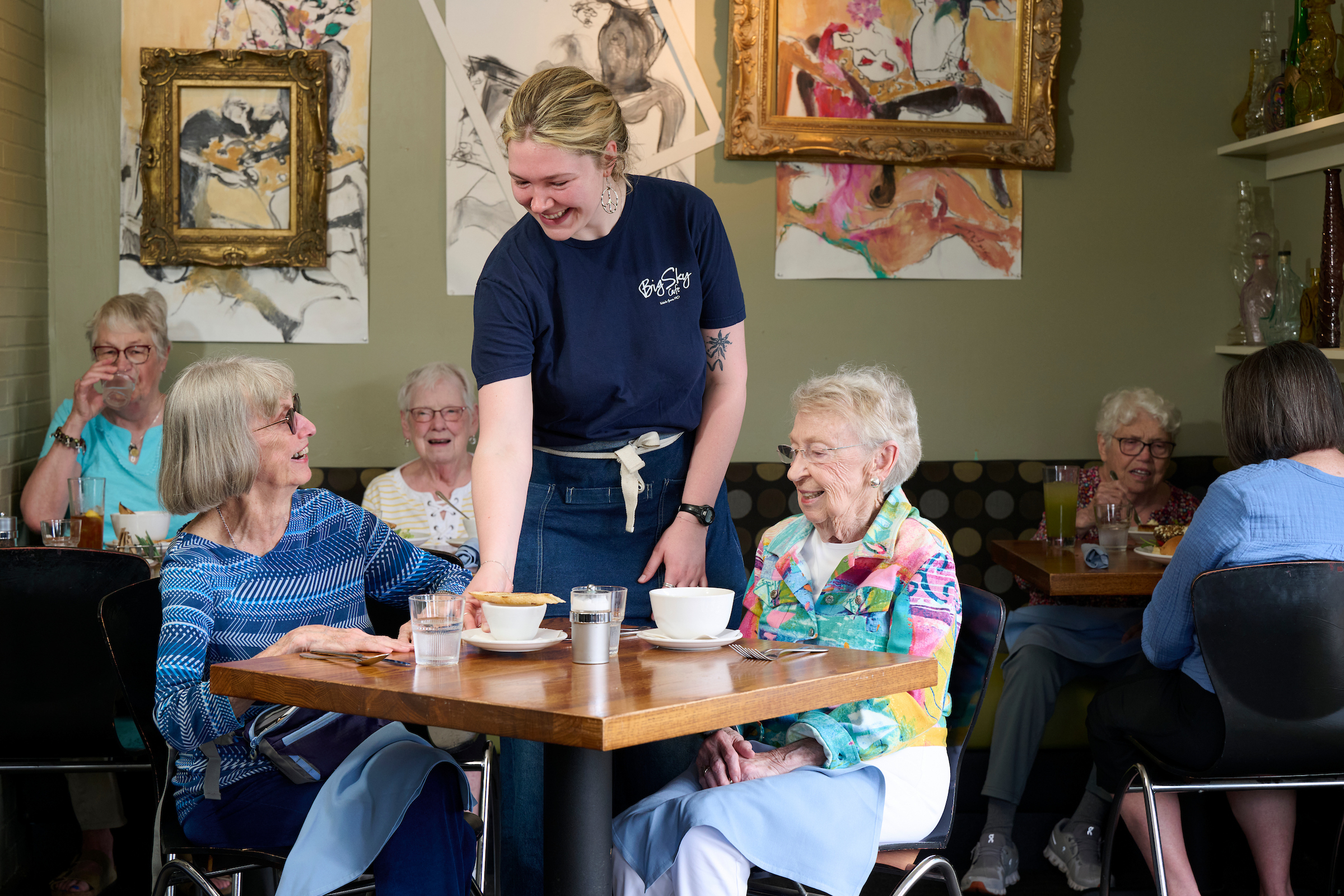Waitress in a navy apron serves dessert to a group of elderly women at a cafe table, with colorful wall art in the background.