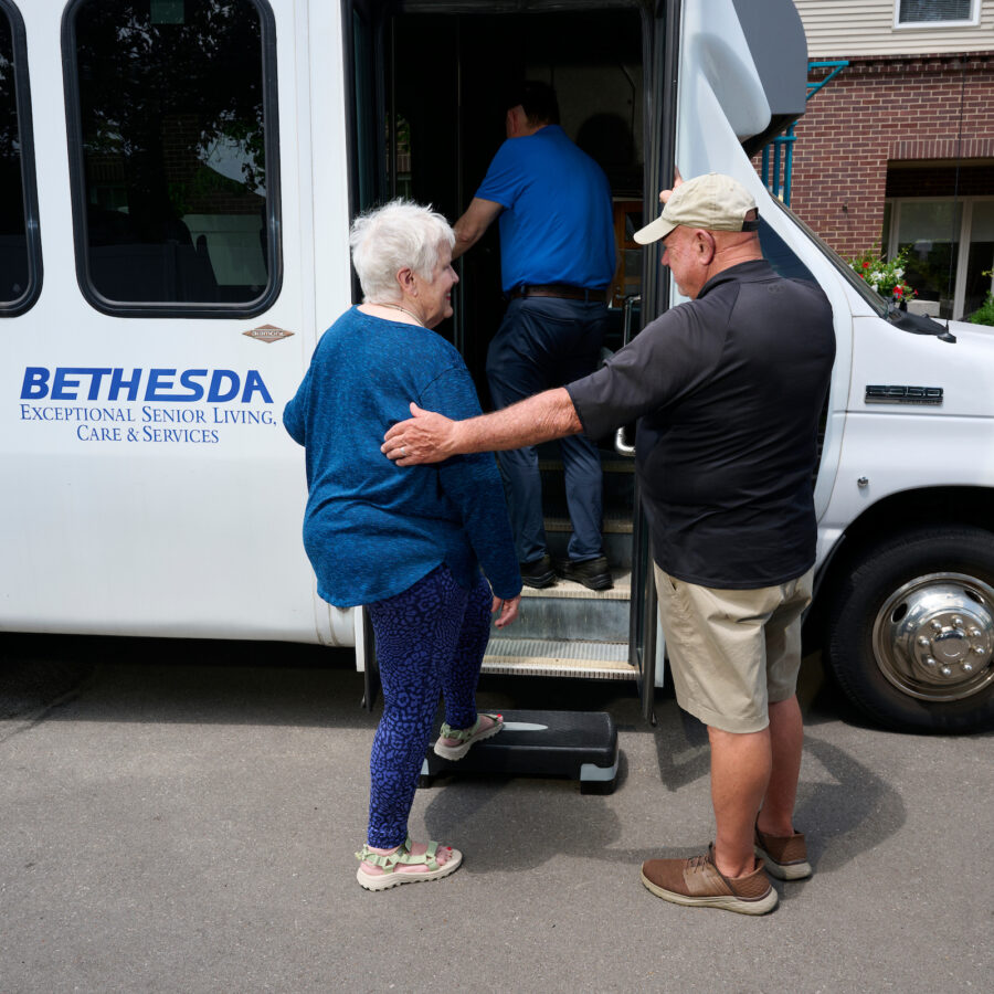 Older adults boarding a white Bethesda shuttle bus as staff assist them at the door.