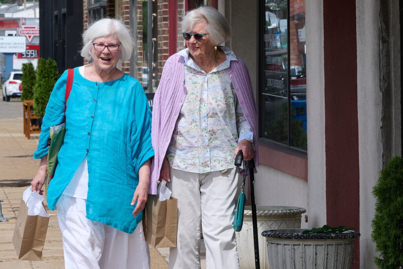 Two elderly women walk together on a sunny sidewalk, carrying shopping bags, one using a cane.