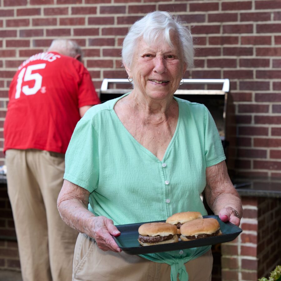 Smiling elderly woman in a mint blouse holds a tray of mini cheeseburgers at a backyard barbecue, brick wall in the background.