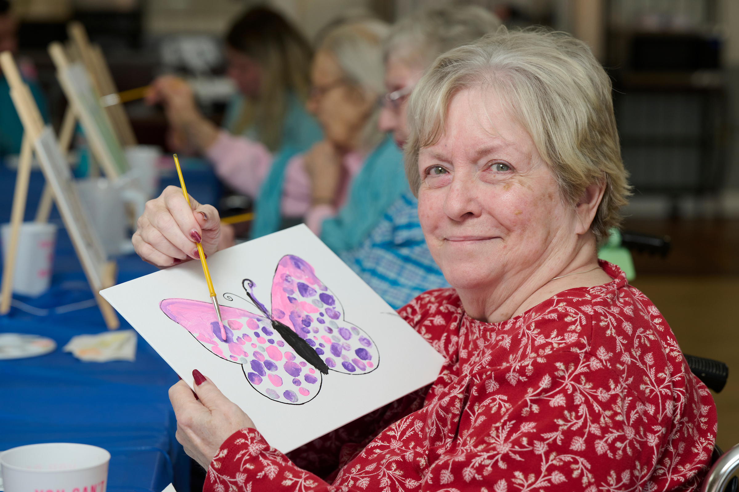 Smiling elderly woman in a red patterned blouse shows a butterfly painting she made, with easels in the background.