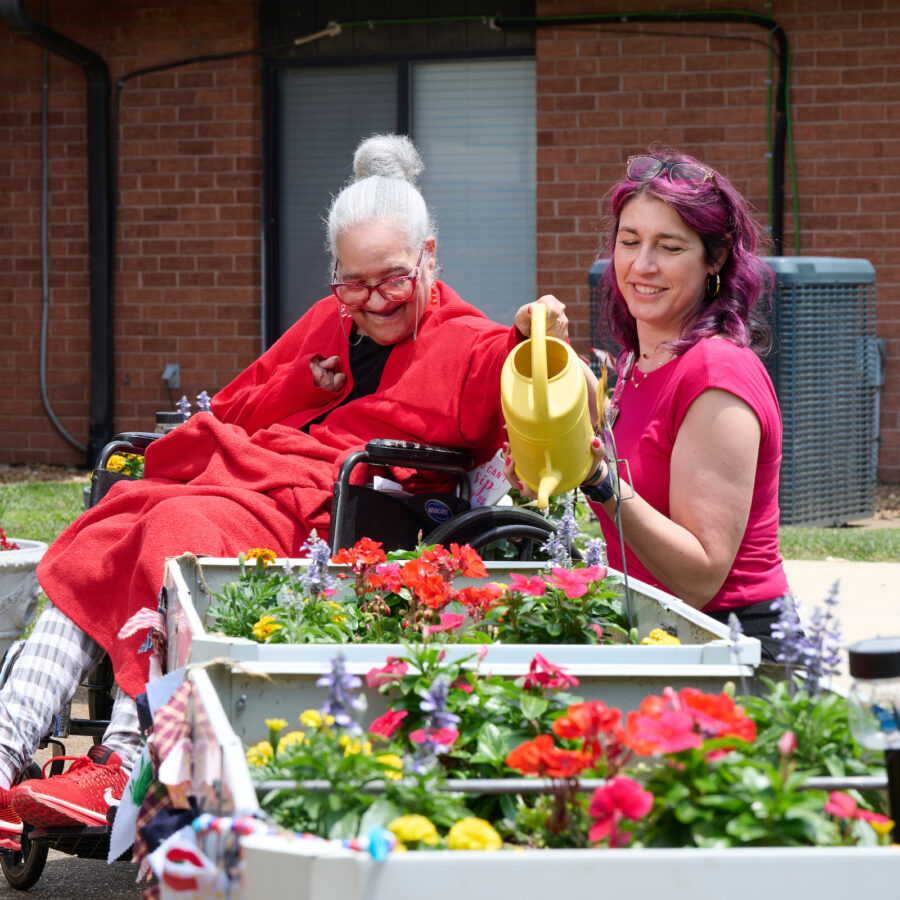 Two women garden outside a brick house: one in a wheelchair with a red blanket waters a flower planter while smiling at her companion.