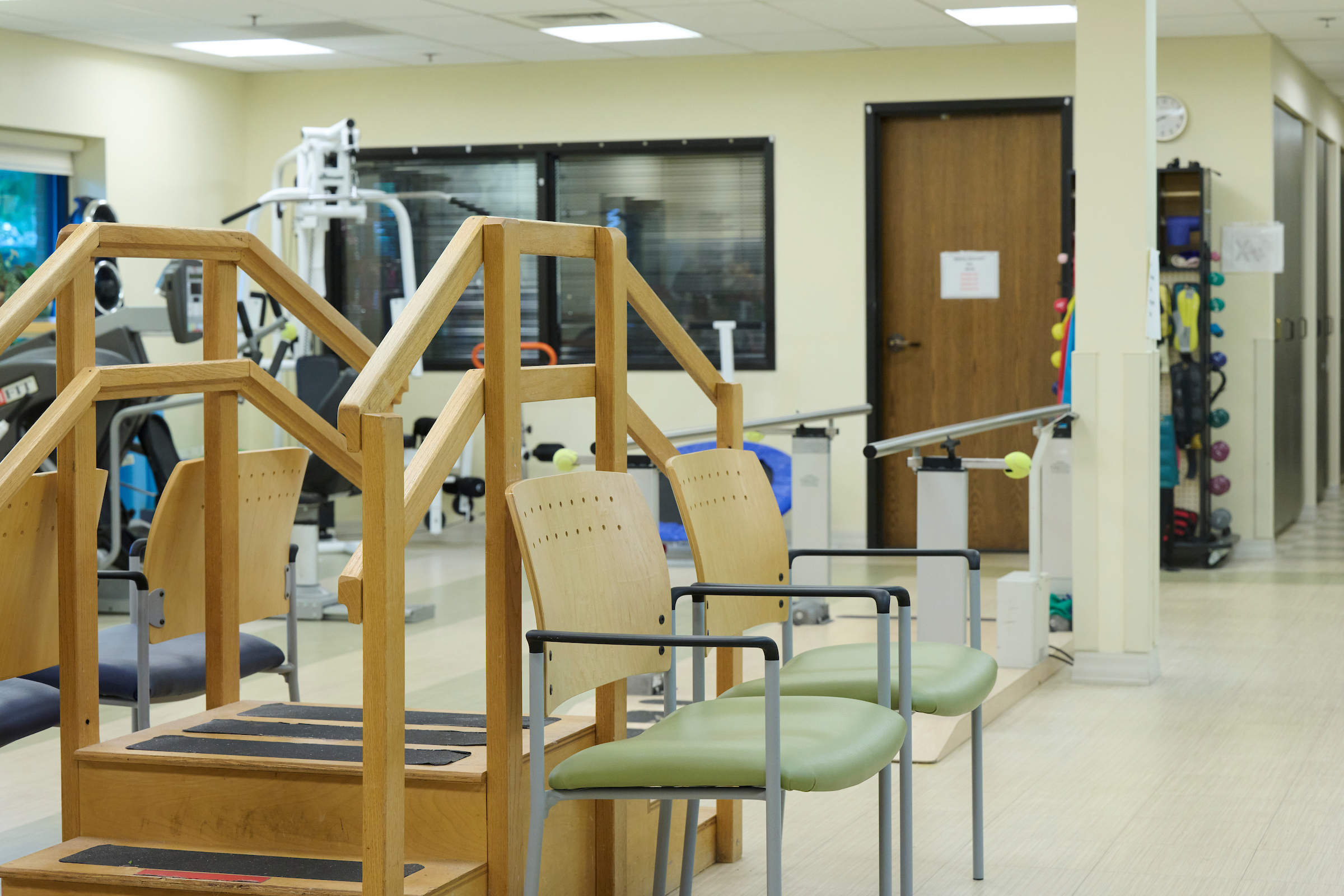 Wooden balance step structure in foreground of a physical therapy room, with green cushioned chairs and exercise equipment in the background.