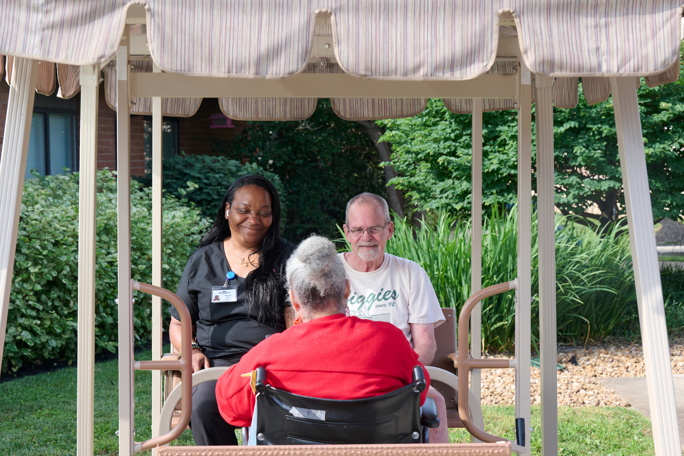 Caregiver and two adults seated on an outdoor wheelchair swing, smiling under a striped canopy.