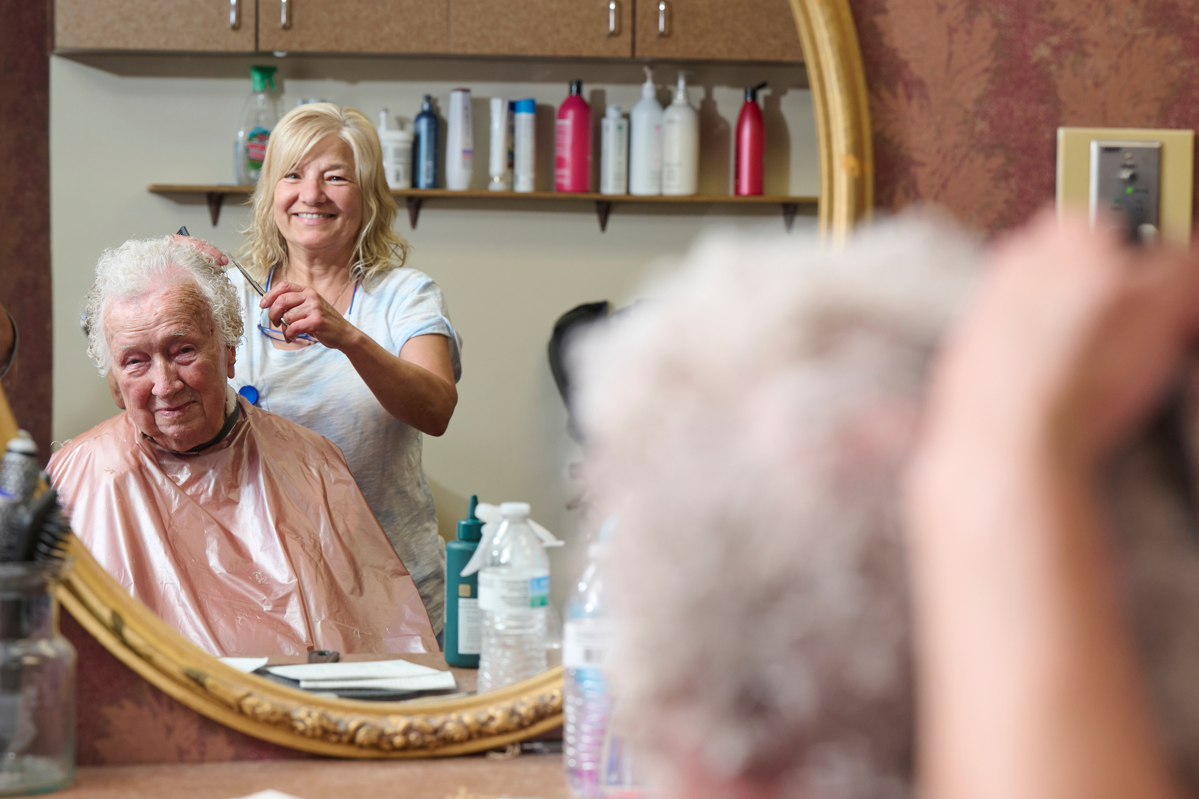 Smiling female hairstylist trims an elderly man's hair as he sits with a pink cape in a salon; round mirror and bottles on a shelf in the background.
