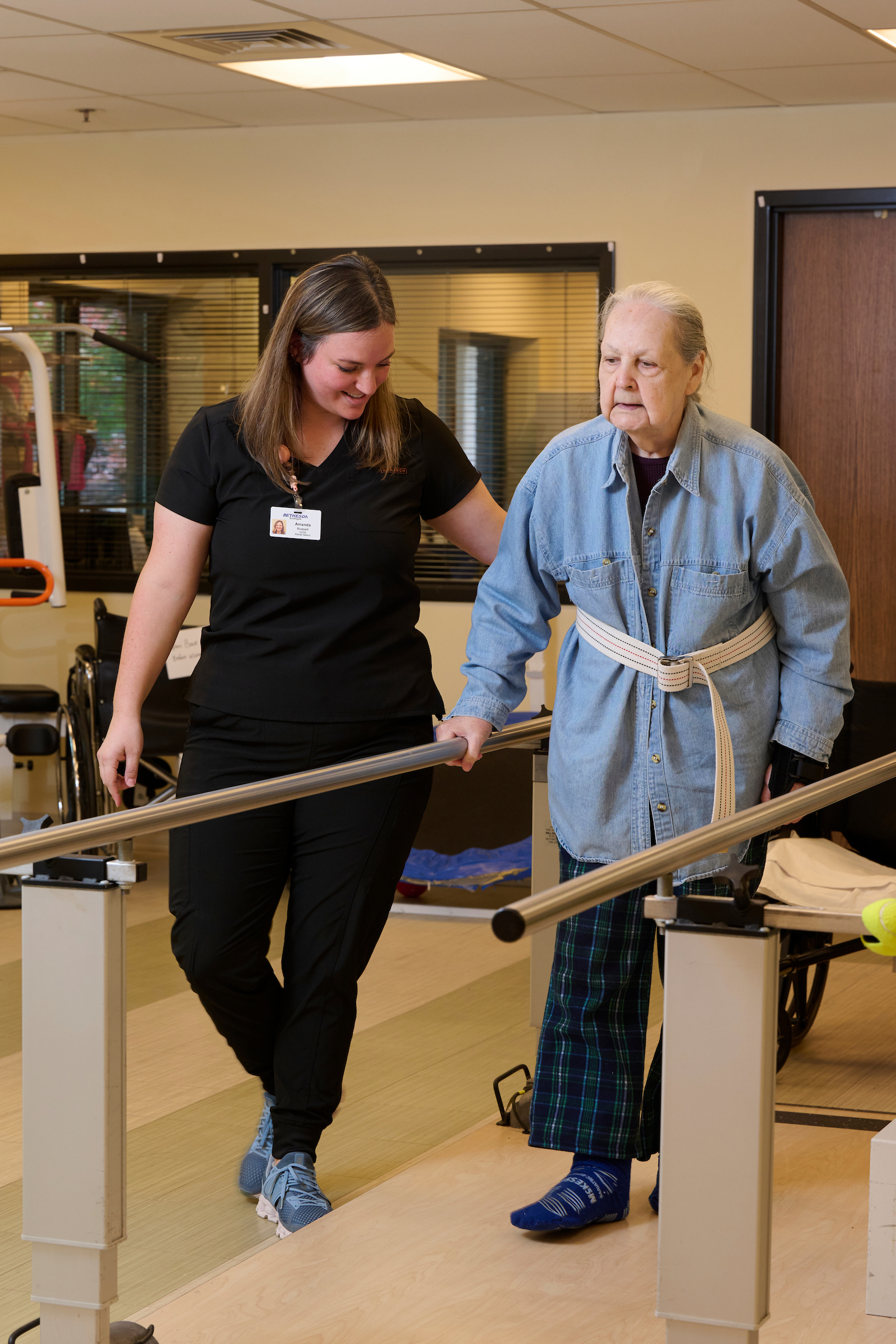 Caregiver assisting an elderly man as they walk along parallel bars in a rehab gym.