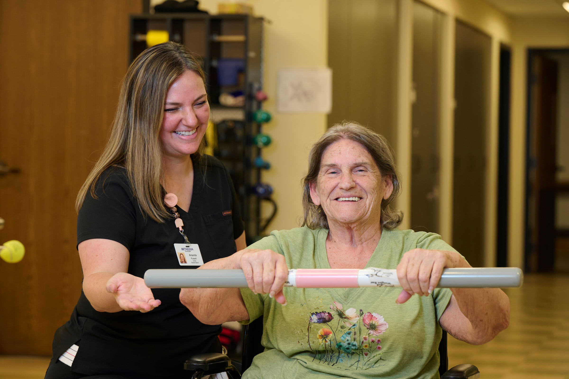Caregiver in black scrubs assists an elderly woman in a wheelchair holding a pink and white bar during exercise in a clinic.