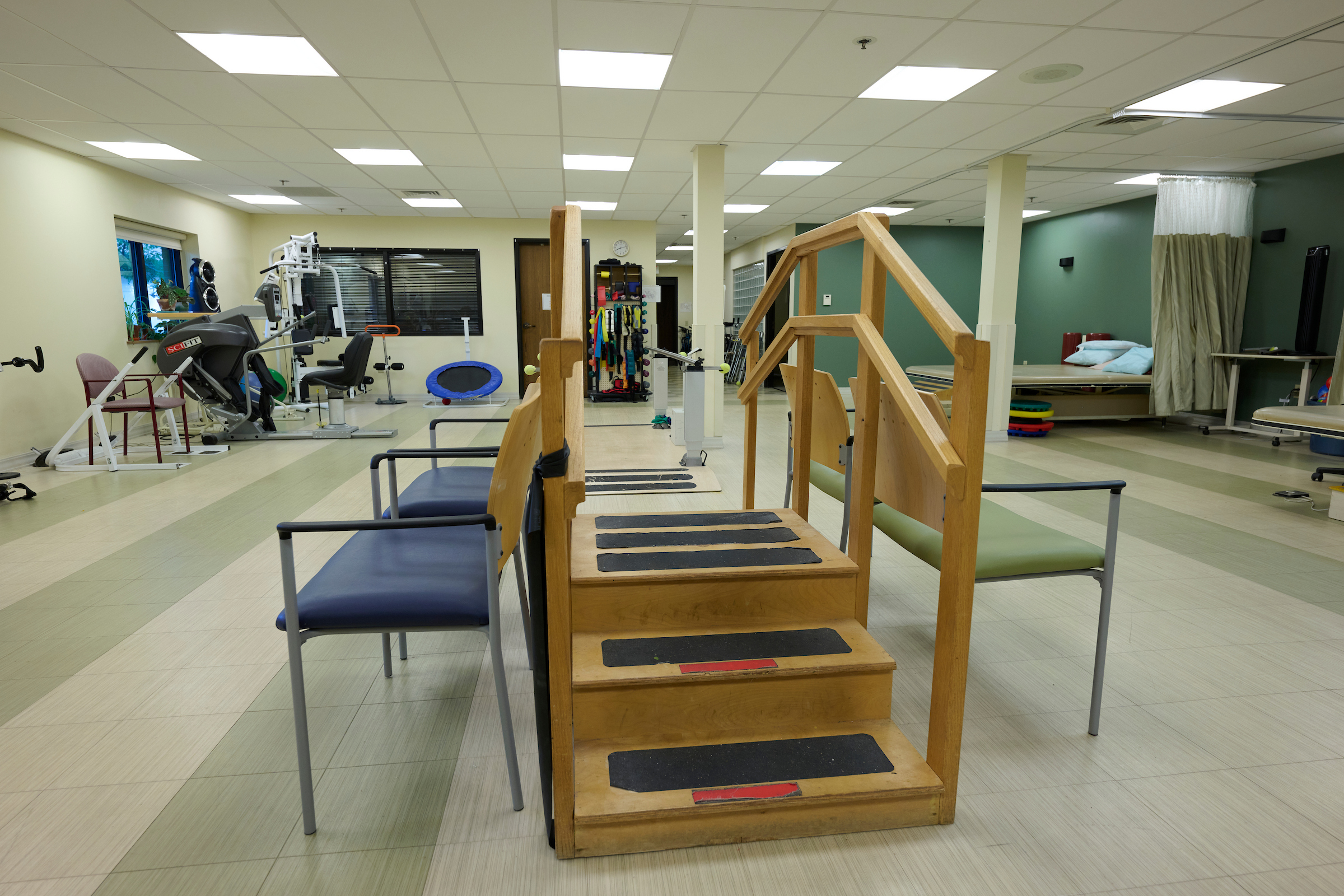 Therapy gym featuring wooden parallel bars and a stepping structure, with exercise equipment along pale walls and a window to the left.