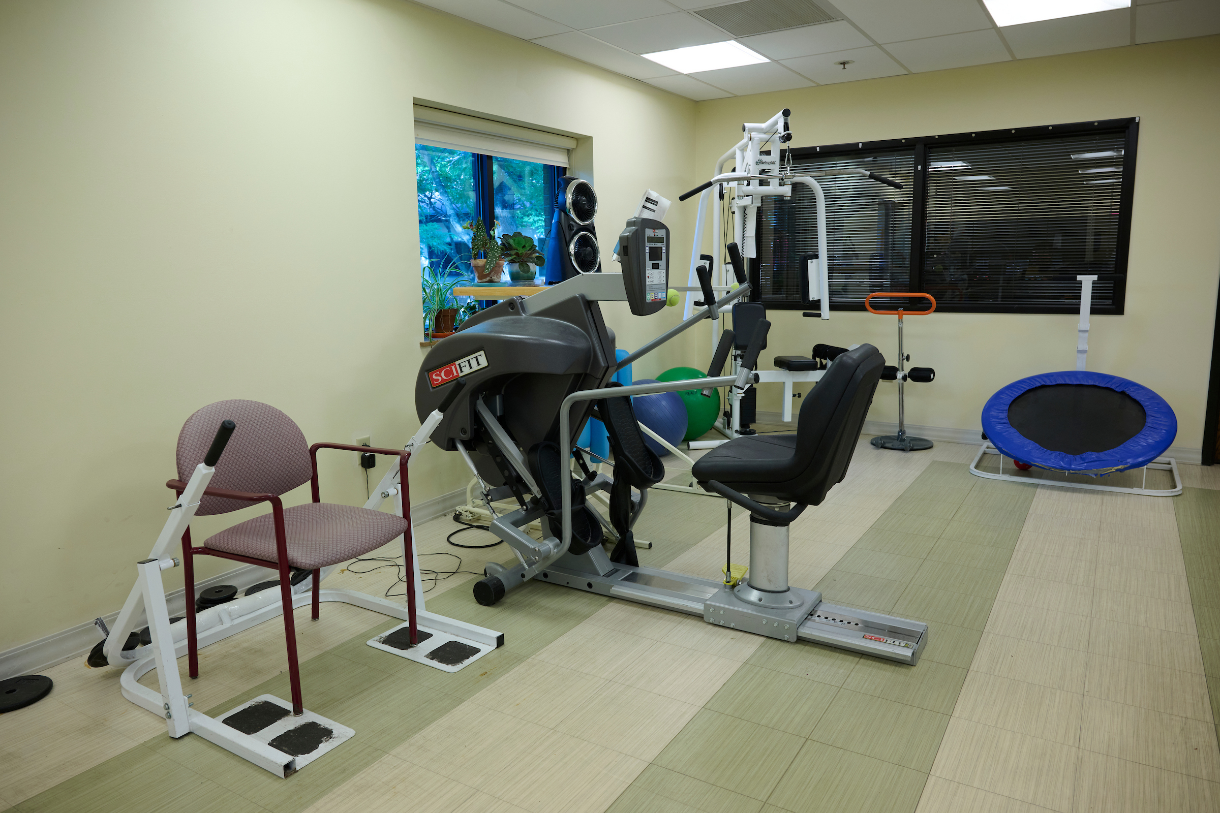 Rehabilitation gym with cardio machines, a weight‑lifting frame, and a blue mini‑trampoline by a window with potted plants.