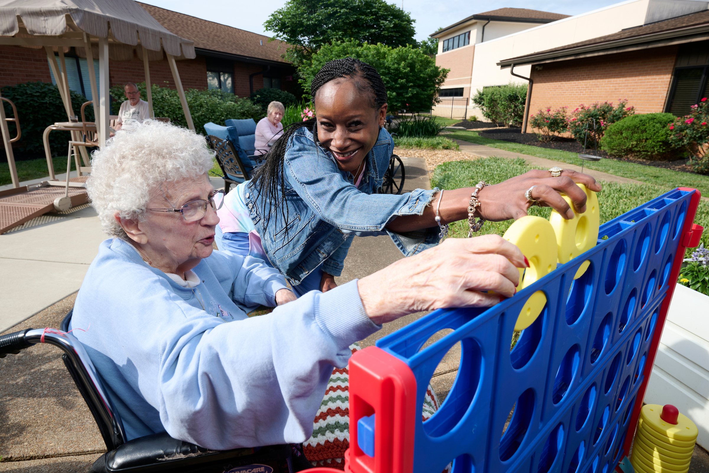 Older woman in a wheelchair reaches toward a colorful play cart while a younger helper supports her outdoors under sunny skies.