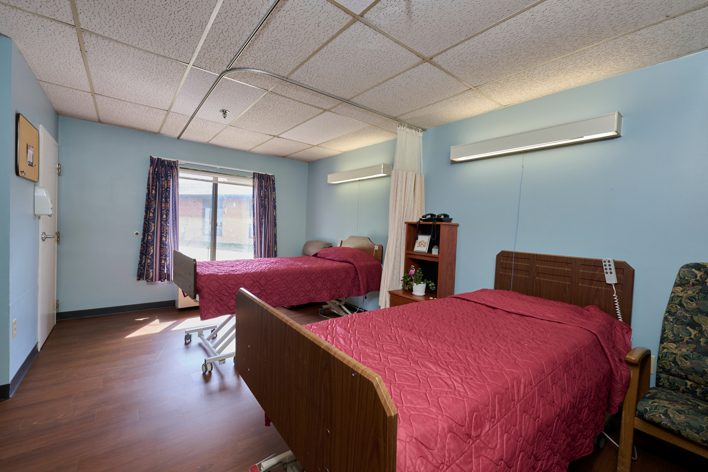 Two red-quilted hospital beds in a blue-walled patient room with a window and patterned curtains.