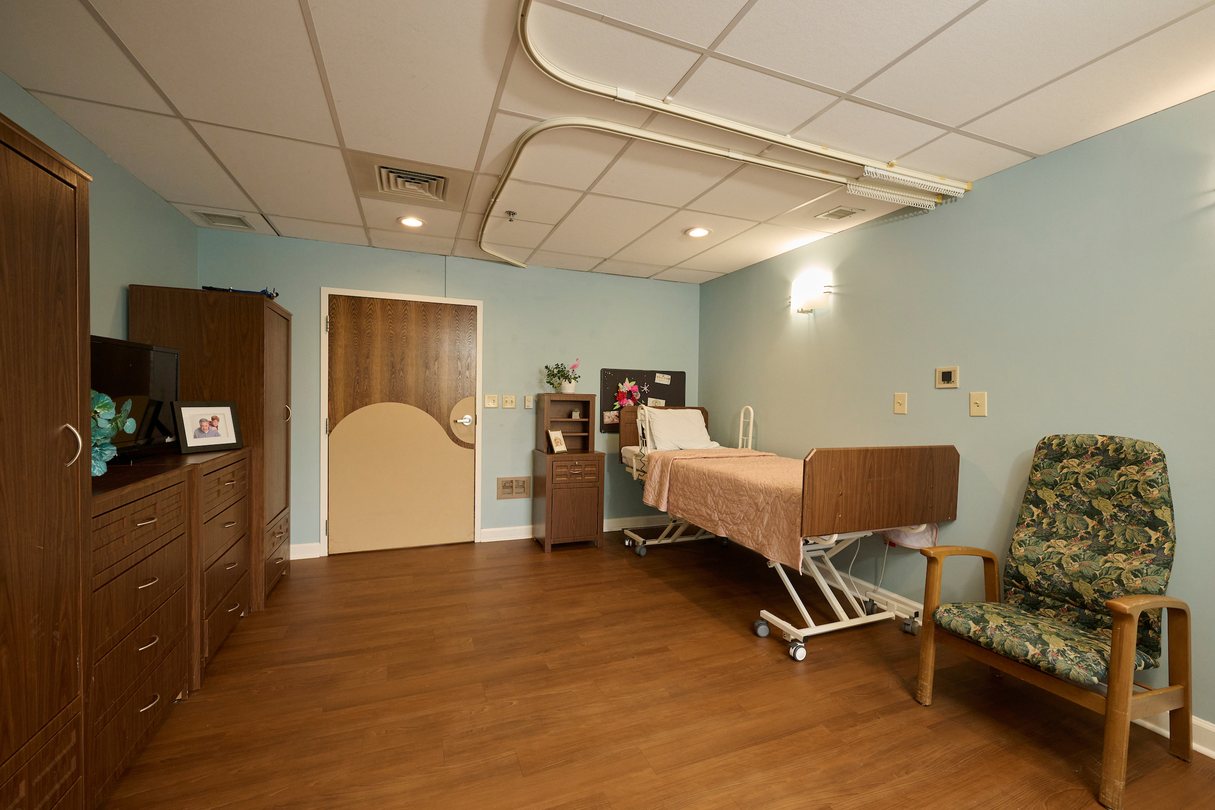 A hospital-style patient room with a mobile adjustable bed and wood furniture lining the left wall, teal walls, and a floral upholstered chair on the right.