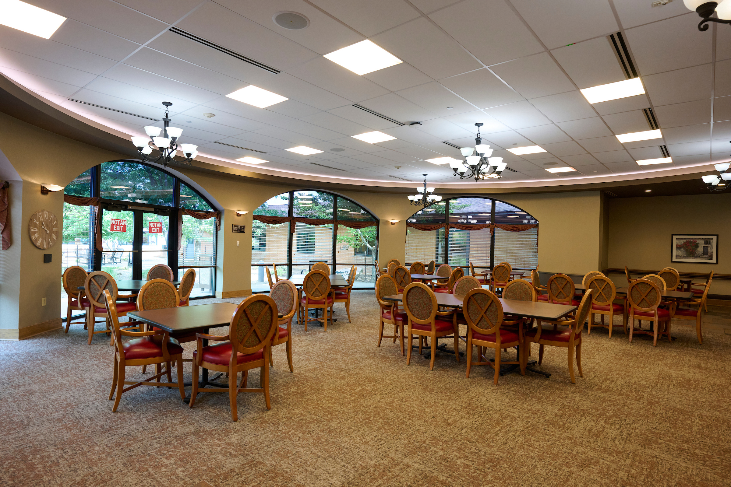 Restaurant-style dining room with numerous round tables and upholstered chairs, arched windows, and chandeliers. Sunlight outside creates a warm atmosphere.