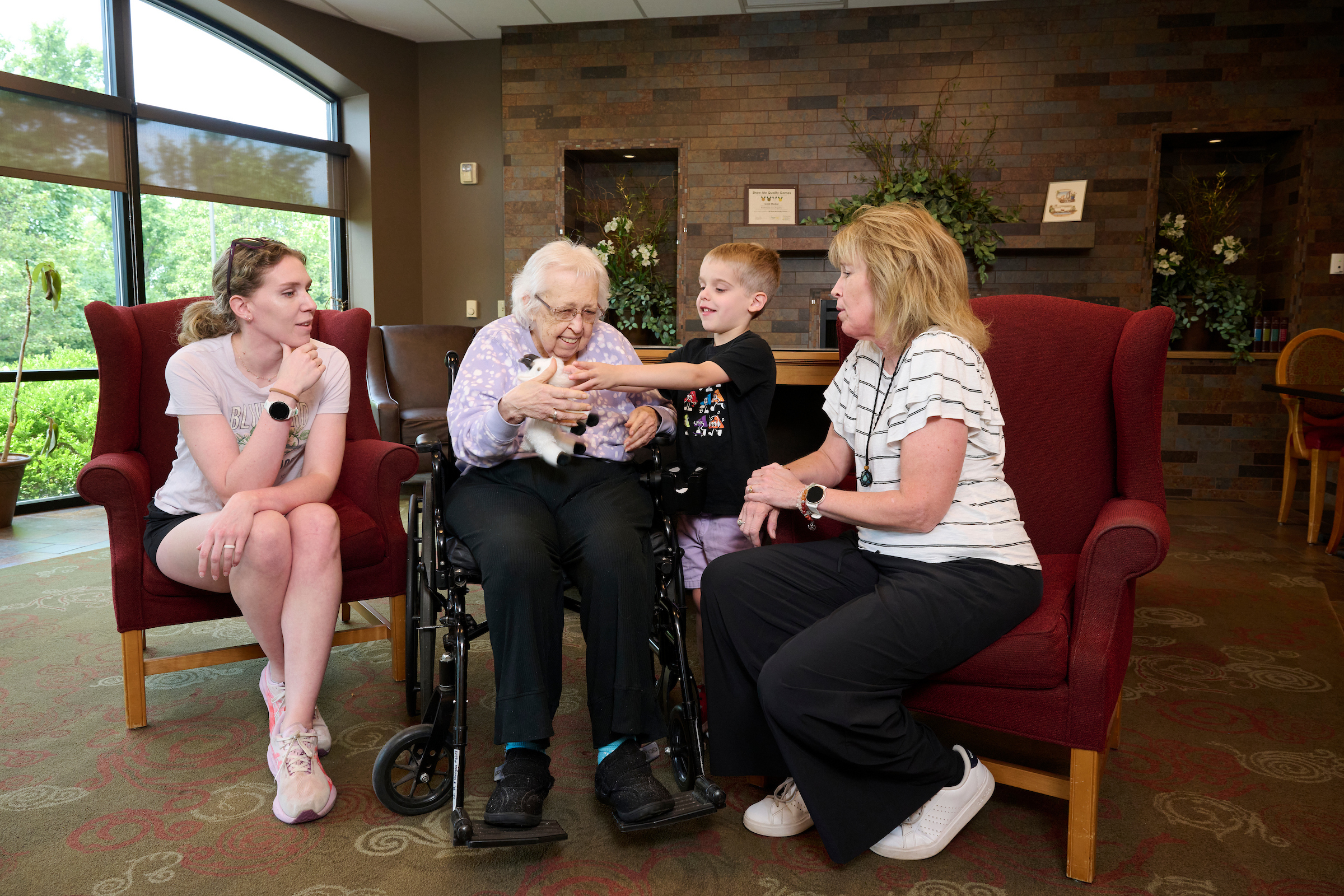 Elderly woman in a wheelchair holds a small black-and-white puppy while a boy and two women sit nearby in a lounge with red chairs and a brick wall backdrop.