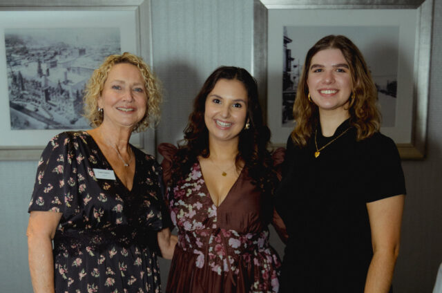 Three smiling women posing together indoors, wearing floral and dark dresses, with framed art/photos on a gray wall behind them.